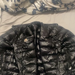 Svart Moncler dunjacka i glansigt material - Säljer en svart dunjacka från Moncler med glansig finish och klassisk quiltad design. Jackan har dragkedjor på sidorna och Moncler-logga på bröstet. Perfekt för kalla dagar när du vill hålla stilen och värmen. Det är M passar L