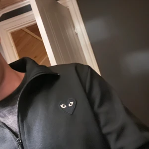 Svart jacka Comme des Garçons tröja - Svart tröja från Comme des Garçons Play med dragkedja och ikoniskt hjärta med ögon på bröstet och ryggen. Jackan har ståkrage, två fickor framtill och är i ett mjukt material. Perfekt för dig som gillar streetwear och stilrena detaljer.