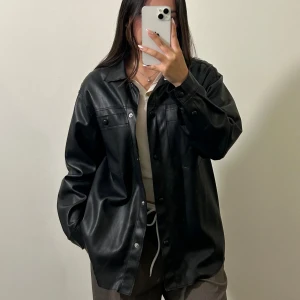 Svart overshirt i skinn från pull & bear  - Säljer en svart overshirt i skinnimitation från Pull & Bear. Jackan har en loose passform, klassisk krage, tryckknappar framtill och två bröstfickor. I superbra skick, skriv för flera bilder 😊.