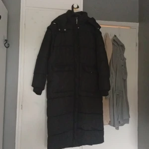 Svart lång pufferjacka från ADPT - Lång svart pufferjacka från ADPT med hög krage, avtagbar huva och stora fickor med tryckknappar. aldrig använd. Pris kan diskuteras