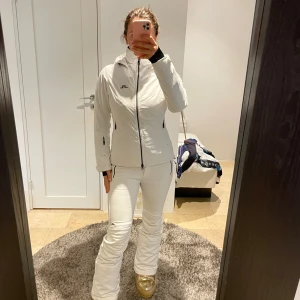 Vit skidjacka från J.Lindeberg - Snygg vit skidjacka från J.Lindeberg med huva, svarta dragkedjor och logga på bröstet. Jackan har figurnära passform, flera fickor med dragkedja och är fodrad för extra värme. Perfekt för vinterns äventyr i backen. Inga defekter förutom den svarta pricken vid en dragkedja, (sista bilden) nypris 4200kr 