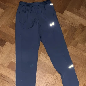 Blå träningsbyxor från Under Armour - Snygga mörkblå träningsbyxor från Under Armour med elastisk midja och reflekterande detaljer. Byxorna har en rak passform och är gjorda i lätt syntetmaterial som andas. Perfekta för löpning eller gymmet, med coolt logotryck på benet.