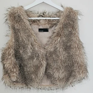 Fuskpälsväst – Vero Moda  - Kort fuskpälsväst från Vero Moda. Har ett litet spänne framtill för att stänga västen. Obs! Ett litet hål i fodret på insidan,inget som syns när den är på. (se bild) Storlek L Bra skick med mindre defekt (se bild)