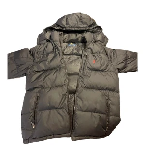 Svart dunjacka från Polo Ralph Lauren - Säljer en svart dunjacka från Polo Ralph Lauren med huva och dragkedja Jackan har två fickor med dragkedja och en röd broderad logga på bröstet. Puffig modell som håller dig varm under kalla dagar. Perfekt för dig som gillar klassisk streetstyle.