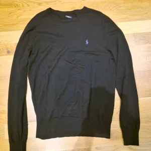 Svart tröja från Ralph Lauren - Klassisk svart tröja från Ralph Lauren med rund halsringning och lila broderad logga på bröstet. Tröjan har lång ärm och ribbade muddar vid ärmslut och nederkant. Perfekt för en stilren och enkel look. Storlek m passar 180, samt ett litet hål på armen som knappast syns