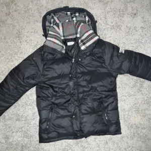 Svart pufferjacka från Burberry - Svart pufferjacka från Burberry med klassiskt rutigt foder i grått, vitt, svart och rött. Jackan har huva, dragkedja och knappar framtill samt två fickor med dragkedja. Stor logopatch på ärmen. Perfekt för kalla dagar och riktigt snygg streetstil . Även kommer passa dig som L,Xl