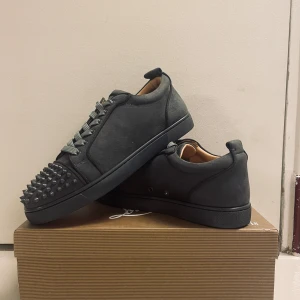 Christian Louboutin grå sneakers med nitar - Svarta sneakers från Christian Louboutin i mocka med coola gråa nitar på tån och klassisk röd sula. Skorna har snörning och rund tå, samt en platt sula. Insidan är i beige skinn med guldfärgad logga. Perfekt för dig som vill sticka ut med lyxig streetstyle.