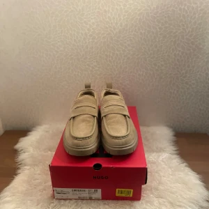 Beige mockaloafers från Hugo - Snygga loafers från Hugo i beige mocka med klassisk pennyloafers-design och grov platt sula. Skorna har rund tå, diskret Hugo-logga på ovansidan och praktisk hälflik. Perfekt för dig som gillar stilren och modern look. Pris går att diskuteras.