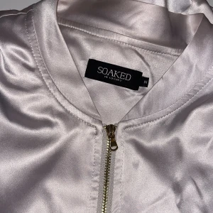 Champagnefärgad satin bomberjacka Soaked - Snygg champagnefärgad bomberjacka från Soaked in Luxury i blankt satinmaterial. Jackan har guldig dragkedja, ribbade muddar och två fickor. Perfekt för dig som vill ha en clean och stilren look med lite extra shine.