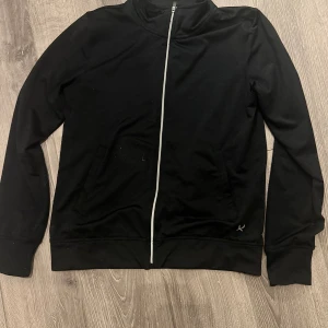 Svart träningsjacka XS - Svart träningsjacka från SOC i storlek XS. Jackan har en stilren design med vit dragkedja framtill, högt krage och två sidofickor. Materialet är mjukt och lätt polyester som andas.