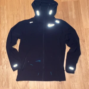 Svart Nike windbreaker - Svart Nike Miller med huva och reflekterande detaljer. Jackan har hel dragkedja framtill, långa ärmar och flera fickor. Perfekt för träning utomhus och har en clean, sportig look med Nike-logga på bröstet.