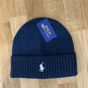 Mörkblå ribbstickad mössa Polo Ralph Lauren - Snygg mörkblå ribbstickad mössa från Polo Ralph Lauren med klassisk broderad logga framtill. Mössan har uppvikt kant och är tillverkad i mjukt material som håller dig varm under kalla dagar. Perfekt accessoar för en stilren look.