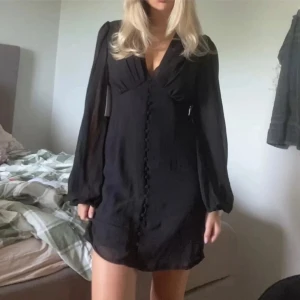 Svart kort klänning med knappar - Supersnygg svart kort klänning med v-ringning och långa, transparenta ärmar. Klänningen har dekorativa knappar framtill och ett lätt, flowy tyg som ger en chic vibe. Perfekt för dig som gillar stilrena och trendiga plagg.