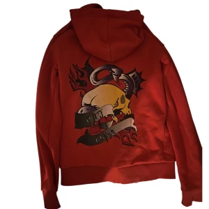 Röd hoodie med döskalletryck och orm - Röd hoodie med dragkedja och huva. På ryggen finns ett stort tryck med döskalle, orm, hjärtan och eldflammor i gult, svart, lila och rött. Tröjan har känguruficka framtill och ribbade muddar. Perfekt för dig som gillar edgy streetstyle.