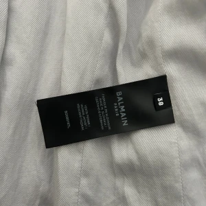 Vit kavaj från Balmain med guldknappar - Snygg vit kavaj från Balmain Paris i storlek 38. Kavajen har markerade axlar, klassisk krage och sex guldiga knappar vid ärmslutet. Fickor med lock och figurnära passform ger en stilren look. Perfekt för dig som vill ha en exklusiv vibe.