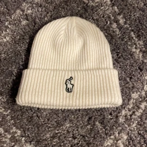 Vit ribbad mössa från Polo Ralph Lauren - Vit ribbstickad mössa från Polo Ralph Lauren med klassisk uppvikt kant och broderad logga framtill. Snygg och enkel design som passar till allt, tillverkad i mjukt stickat material som håller dig varm under kalla dagar.