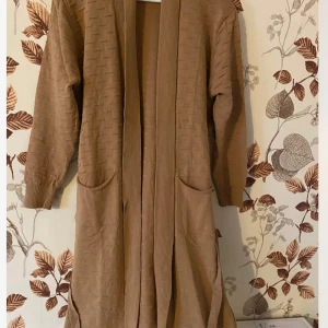 Beige lång kofta med fickor - Lång beige kofta med snyggt stickat mönster och två stora fickor framtill. Koftan har ribbade muddar och bred ribbad kant längs öppningen. Perfekt att slänga på sig över en t-shirt eller topp för en chill look.