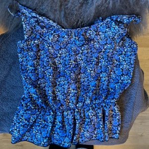 Blommig blå blus från Bik Bok XS - Supersöt blommig blus från Bik Bok i 💙 och 🖤. Modellen har smala axelband med knyt, volangkant upptill och resår i midjan för en snygg passform. Perfekt för dig som gillar färg och mönster!