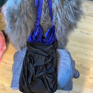 Svart och blå kortklänning med omlott - Säljer en snygggg kortklänning i svart med blå omlottdetaljer upptill. Klänningen har axelbandslös design med draperad effekt över bysten och smala band som knyts i nacken. 