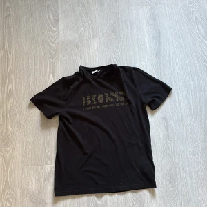 Svart t-shirt från Hugo Boss - Svart t-shirt från Hugo Boss med guldigt BOSS-tryck framtill. Klassisk rund hals och korta ärmar. Skön och stilren modell i mjuk bomull, perfekt för en clean och snygg look.