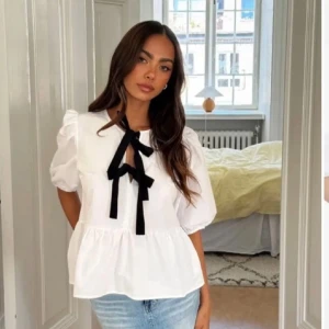 Vit blus med svarta rosetter från Bubbleroom - Supersöt vit blus från Bubbleroom med puffiga korta ärmar och två svarta rosetter framtill. Blusen har en lätt volang nertill. Perfekt för dig som gillar en trendig och söt stil!