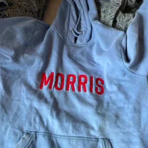 Ljusblå hoodie från Morris XS - Ljusblå hoodie från Morris med röd broderad logga framtill. Tröjan har huva med snörning och en klassisk känguruficka. Tillverkad i mjuk bomull och polyesterblandning, perfekt för chill dagar. Snygg och enkel att matcha med jeans eller joggers.
