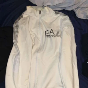 Vit EA7 vindjacka med dragkedja - Snygg vit vindjacka från EA7 Emporio Armani med hel dragkedja framtill och svarta ränder på axlarna. Jackan har hög krage och EA7-logga på bröstet. Perfekt för dig som gillar sportig och clean stil.