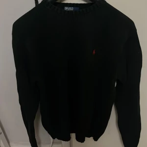 Svart stickad tröja Polo Ralph Lauren - Klassisk svart stickad tröja från Polo Ralph Lauren med ett rött broderat Ralph Lauren logga på bröstet. Tröjan har rund halsringning, ribbade muddar och lång ärm. Perfekt för att matcha med jeans eller chinos. Kan gå ner i pris vid snabb affär