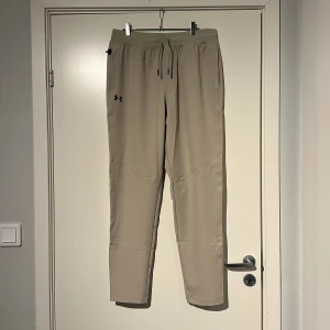 Beige tapered byxor från Under Armour - Snygga beige tapered byxor från Under Armour med elastisk midja och dragsko. Byxorna har en sportig look, sidofickor och diskret logga på låret. Perfekta för en chill vardagsstil eller träning. Tillverkade i mjukt och lätt syntetmaterial för maximal komfort.