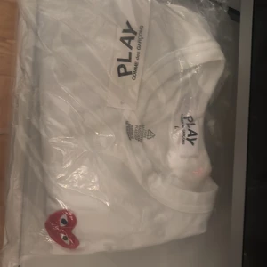 Vit t-shirt Comme des Garçons Play - Vit t-shirt från Comme des Garçons Play med det ikoniska röda hjärtat med ögon broderat på bröstet. Klassisk passform och rund hals. Perfekt för dig som gillar stilrena och trendiga plagg med en twist.