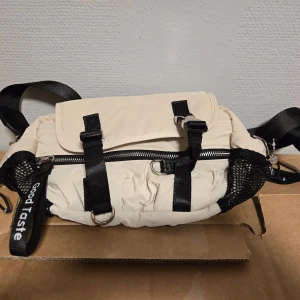 Beige axelväska från Zara - Cool beige axelväska från Zara med svarta detaljer, justerbar rem och flera fack. Väskan har dragkedja, spännen och meshfickor på sidorna. Snygg och praktisk för dig som vill ha en trendig och funktionell väska till vardagen.