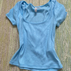 Ljusblå topp från H&M Divided - Säljer en ljusblå topp från H&M Divided med korta ärmar och rundad, djup urringning. Toppen har diskret hålmönster och är i mjuk bomull, perfekt för en chill och fräsch look.