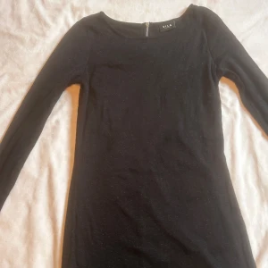 Svart långärmad klänning från Vila - En stilren svart klänning med glitter från Vila med lång ärm och rund halsringning. Klänningen har en smickrande, figurnära passform och är tillverkad i ett mjukt material som sitter bekvämt. Perfekt till nyår. 