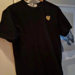 Svart Comme des Garçons Play t-shirt - Svart t-shirt från Comme des Garçons Play i storlek L. Klassisk rund hals och korta ärmar. På bröstet sitter ett broderat guldigt hjärta med ögon, som är ett ikoniskt motiv för märket. Tillverkad i mjuk bomull för skön känsla och snygg passform.