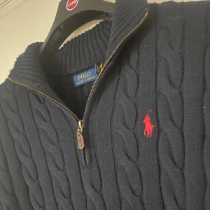 Ralph Lauren Kabelstickad Half Zip - Klassisk kabelstickad tröja från Polo Ralph Lauren med röd logga på bröstet. I princip ny – endast använd en gång och tvättad en gång. Säljes eftersom den är för stor för mig. ✅ Äkta vara (skannbar QR-kod) ✅ Marinblå med röd logga ✅ I princip Nytt skick