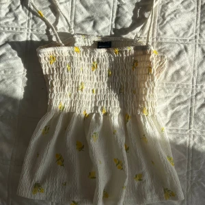 Vit blommig linnetopp från Gina Tricot - Supersöt vit linnetopp från Gina Tricot med smockad överdel och tunna axelband. Toppen har små gula blommor och en lätt volang nertill. Perfekt för varma dagar och ger en fräsch sommarkänsla.