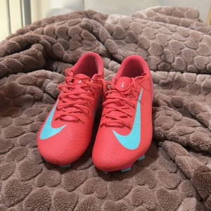 45.5 Nike Zoom Vapor 15 fotbollsskor rosa - Nike Zoom Vapor 15 Academy FG/MG fotbollsskor i en riktigt snygg rosa färg med turkosa detaljer och logga. Skorna har snörning, platt sula med dobbar och är gjorda i syntetmaterial. Perfekta för dig som vill sticka ut på planen.