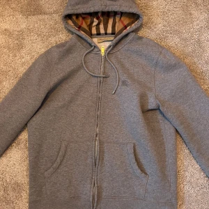 Grå hoodie från Burberry  - Grå hoodie från Burberry med klassiskt rutigt foder i huvan och broderad logga på bröstet. Tröjan har hel dragkedja, snörning i huvan och två fickor framtill. Tillverkad i mjuk bomullsblandning, perfekt för kyliga dagar.