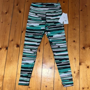 Mönstrade leggings från Röhnisch,storlek S 7/8 - Snygga leggings från Röhnisch i storlek S med coolt randigt mönster i svart, grönt, vitt och grått. Hög midja och tight passform, perfekta för träning. Tillverkade i stretchigt syntetmaterial för maximal komfort.