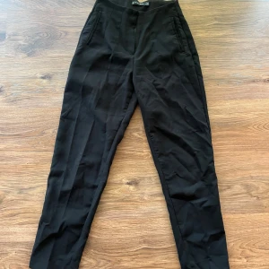 Svarta kostymbyxor  - Svarta kostymbyxor från Amisu i klassisk rak modell med hög midja. Byxorna har diskreta fickor framtill och är perfekta för en clean och stilren look. Materialet känns mjukt och följsamt, vilket gör dem bekväma att bära hela dagen.