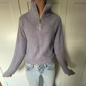 Ljuslila stickad tröja från Kappahl - Supermysig ljuslila stickad tröja från Kappahl med hög krage och half zip-dragkedja. Tröjan har ribbade muddar och en loose passform som ger en chill vibe. Perfekt för kyliga dagar när du vill vara både bekväm och snygg.