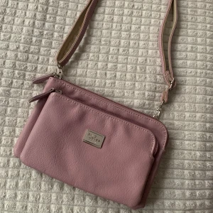 Lila axelremsväska - Liten lila/rosa väska med två stora fack med dragkedja och ett innerfack. Avtagbart band på väskan. Har silver detaljer. En fin väska som är oanvänd.