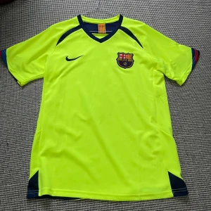 Barcelona Ronaldinho 10 Nike fotbollströja - Säljer en ikonisk Barcelona fotbollströja från Nike med Ronaldinho och nummer 10 på ryggen. Tröjan är neongul med mörkblå detaljer, klubbmärke på bröstet och klassiska ränder i nacken. Tillverkad i lätt och ventilerande polyester, perfekt för match eller träning.