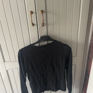 Svart långärmad topp från H&M - Svart långärmad topp från H&M med strukturerad yta och rund halsringning. Toppens material är lätt och stretchigt, vilket ger en bekväm passform. Perfekt att styla med jeans eller kjol för en cool look.