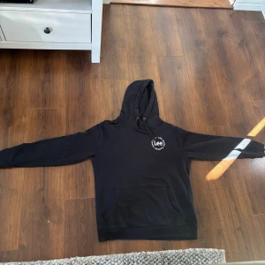 Svart hoodie från Lee med tryck - Svart hoodie från Lee med vit logga på bröstet. Klassisk modell med huva och dragsko, samt stor känguruficka framtill. Mjuk insida och ribbade muddar vid ärmslut och nederkant. Perfekt för chill dagar eller när du vill ha en enkel streetstyle.