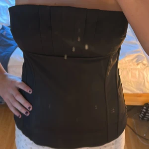 Svart axelbandslös tubtopp i satin - Snygg svart tubtopp i satin med axelbandslös design och stilren form. Toppen har en bandeau-silhuett och diskret söm framtill som ger extra struktur. Perfekt för dig som gillar minimalistisk och elegant stil.