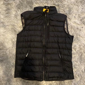 Svart dunväst från Parajumpers - Svart quiltad dunväst från Parajumpers med hög krage och dragkedja framtill. Västen har två snedställda fickor med dragkedja och en lätt, sportig känsla. Perfekt för lager-på-lager och har klassisk Parajumpers-logga i nacken.