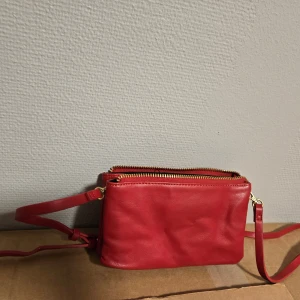 Röd axelväska i skinn med dragkedja - Snygg röd axelväska i skinn med guldfärgade dragkedjor och flera fack. Väskan har en smidig och kompakt design med justerbar axelrem, perfekt för att hålla ordning på dina grejer. Insidan är svart och har praktiska fack för småsaker.