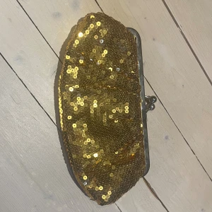 Guldig paljettkuvertväska - Snygg kuvertväska täckt med glittrande guldiga paljetter och metallram med klassiskt lås. Insidan är fodrad i beige tyg och har en liten innerficka. Perfekt för att lyfta din outfit med extra bling.