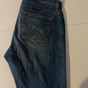 Wrangler Q-Baby blå jeans bootcut - Säljer ett par blå Wrangler Q-Baby jeans med klassisk W-söm på bakfickorna och snygga detaljer i midjan. Modellen har bootcut och normal passform, tillverkad i slitstarkt jeansmaterial med dragkedja och knapp framtill.